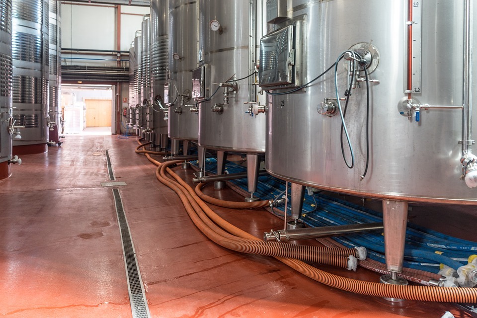 Interior de uma vinícola com tanques de aço inoxidável e mangueiras para fermentação do vinho.